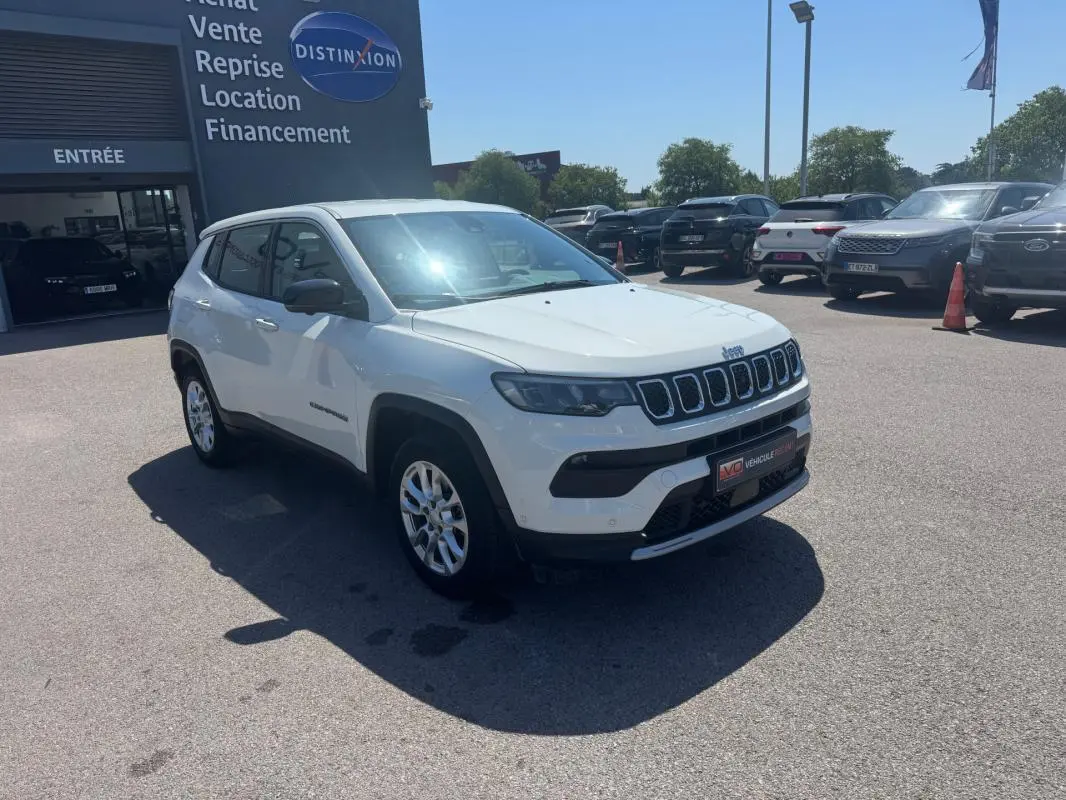 Jeep Compass blanc en 3/4 avant droit, stationné devant un bâtiment commercial sous un ciel dégagé.