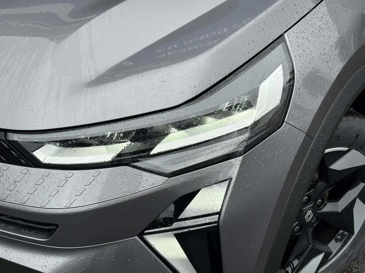 Gros plan sur le phare avant droit et la jante noire du Renault Captur Gris Cassiopée 2025 sous la pluie.