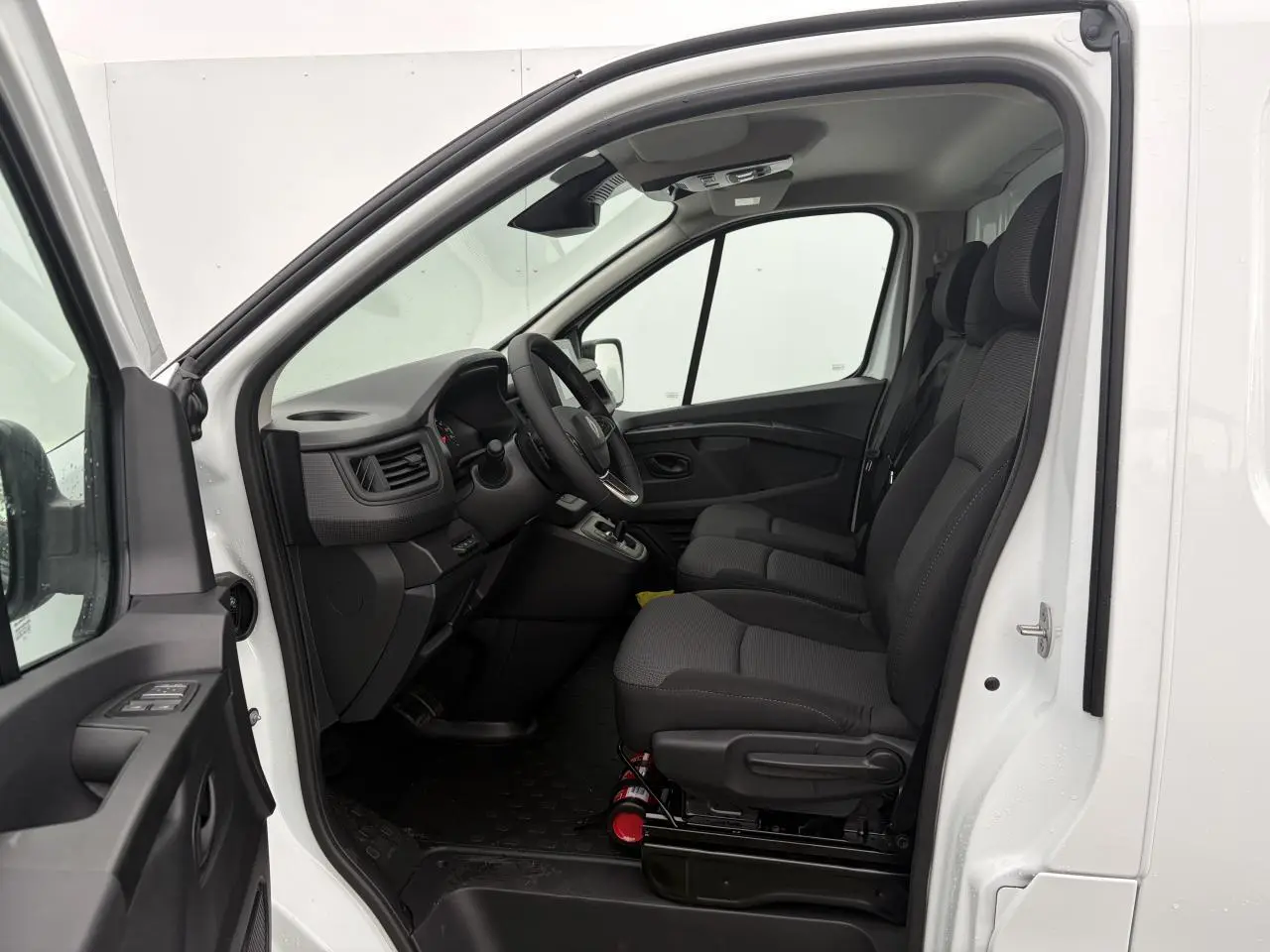 Vue intérieure côté conducteur du Renault Trafic Fourgon blanc glacier, montrant les sièges avant en tissu et le tableau de bord.