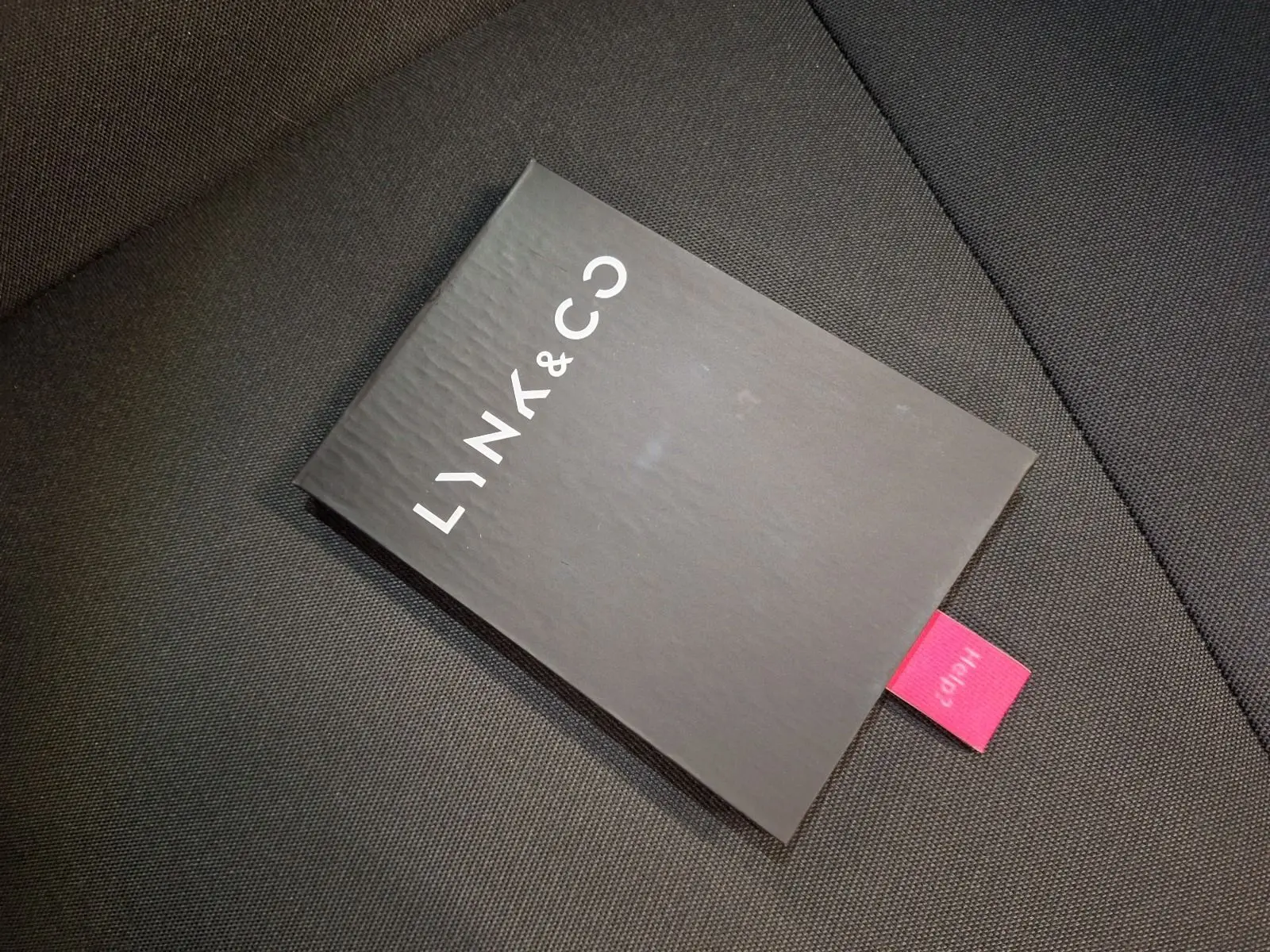 Manuel d'utilisation noir Lynk & Co posé sur un siège en tissu gris foncé, vue en plongée rapprochée.