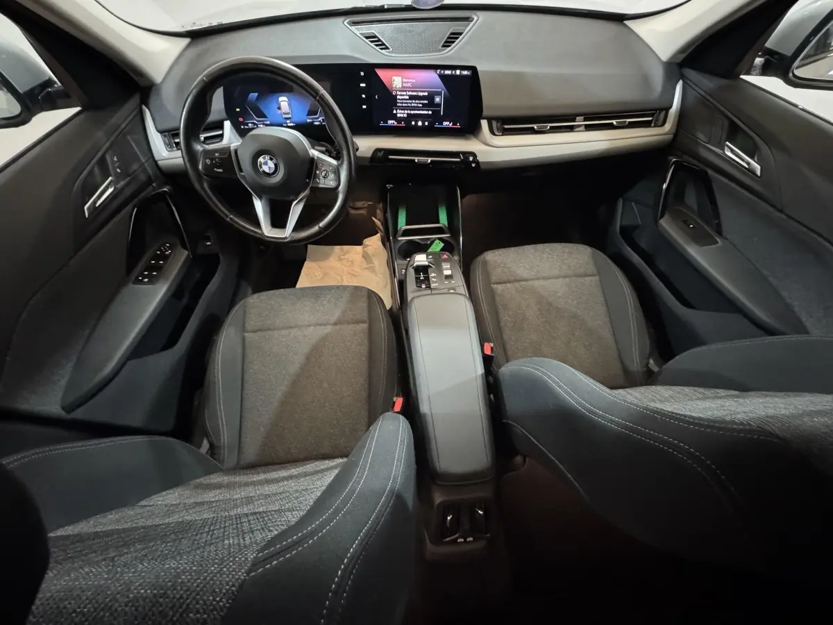 Vue intérieure avant du BMW X1 2024 gris foncé, avec tableau de bord digital et sièges tissu gris.