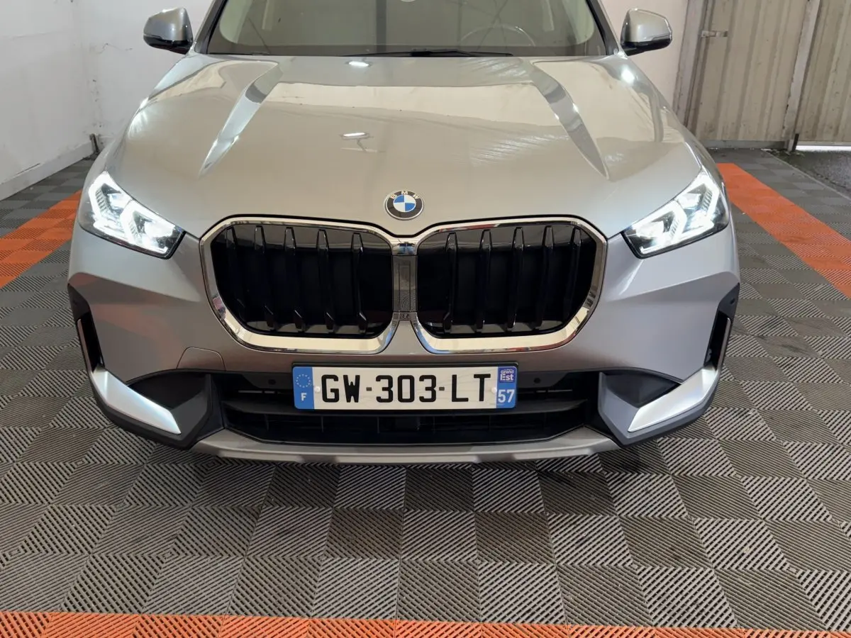Vue frontale d’un BMW X1 gris foncé 2024 avec calandre noire et phares LED allumés dans un garage.