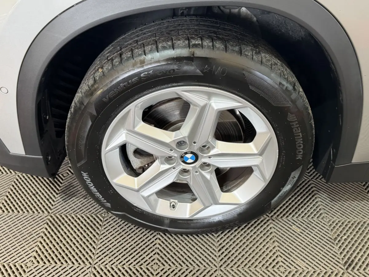 Gros plan sur la roue avant droite avec jante argentée et logo BMW, partie de la carrosserie gris foncé visible.