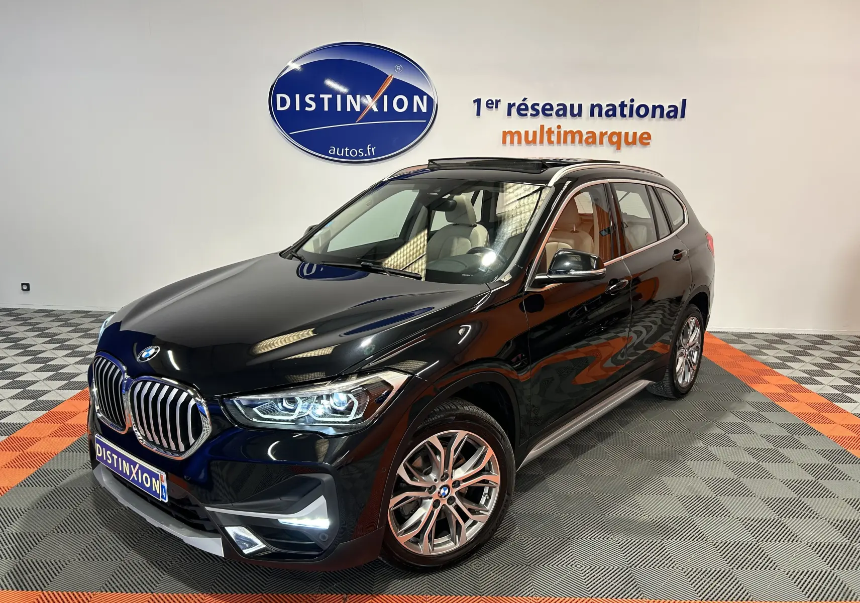 BMW X1 noir en 3/4 avant droit avec toit ouvrant et barres de toit neuves visibles en intérieur showroom.