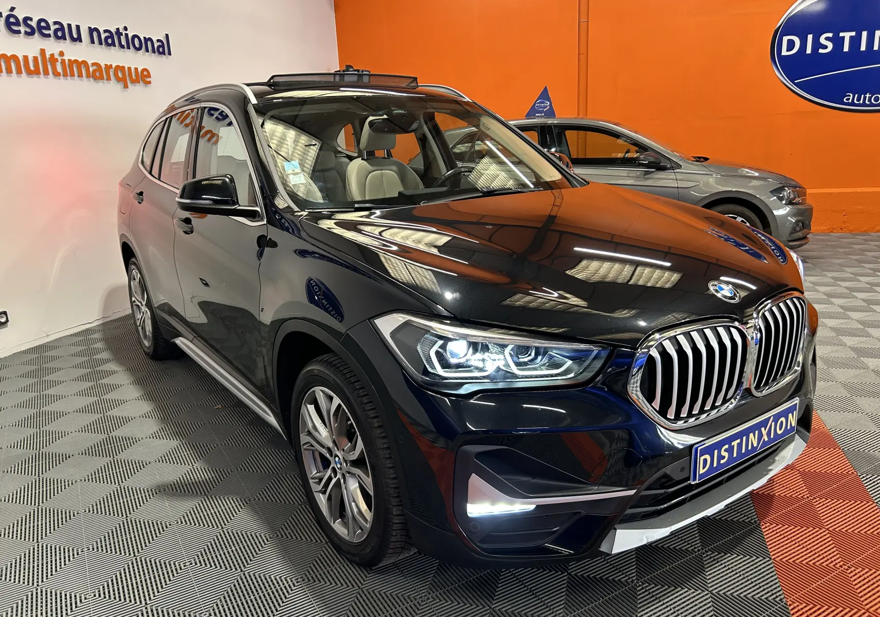BMW X1 sDrive18iA noir en 3/4 avant droit, phares allumés et barres de toit visibles en intérieur showroom.
