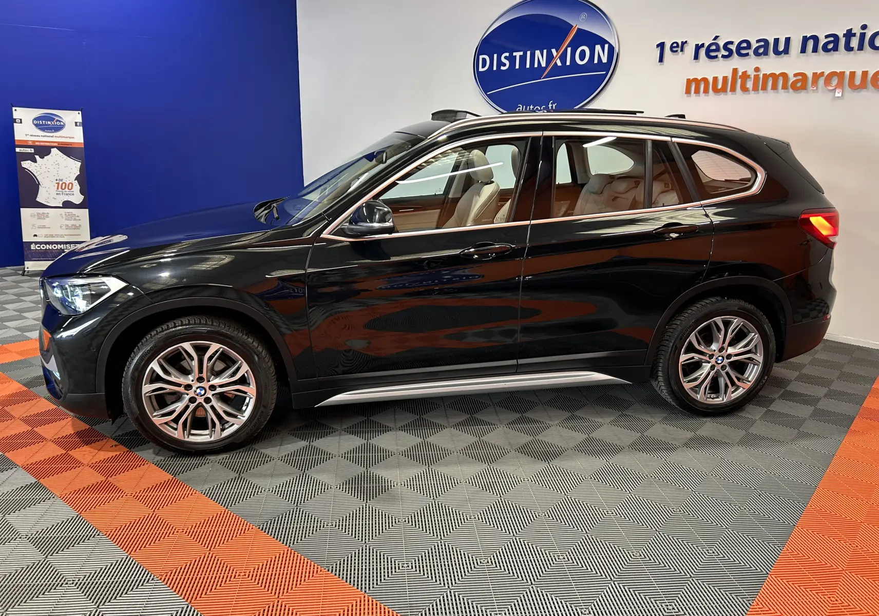 BMW X1 sDrive18iA noir vue de profil côté gauche en intérieur avec toit ouvrant et barres de toit neuves visibles.