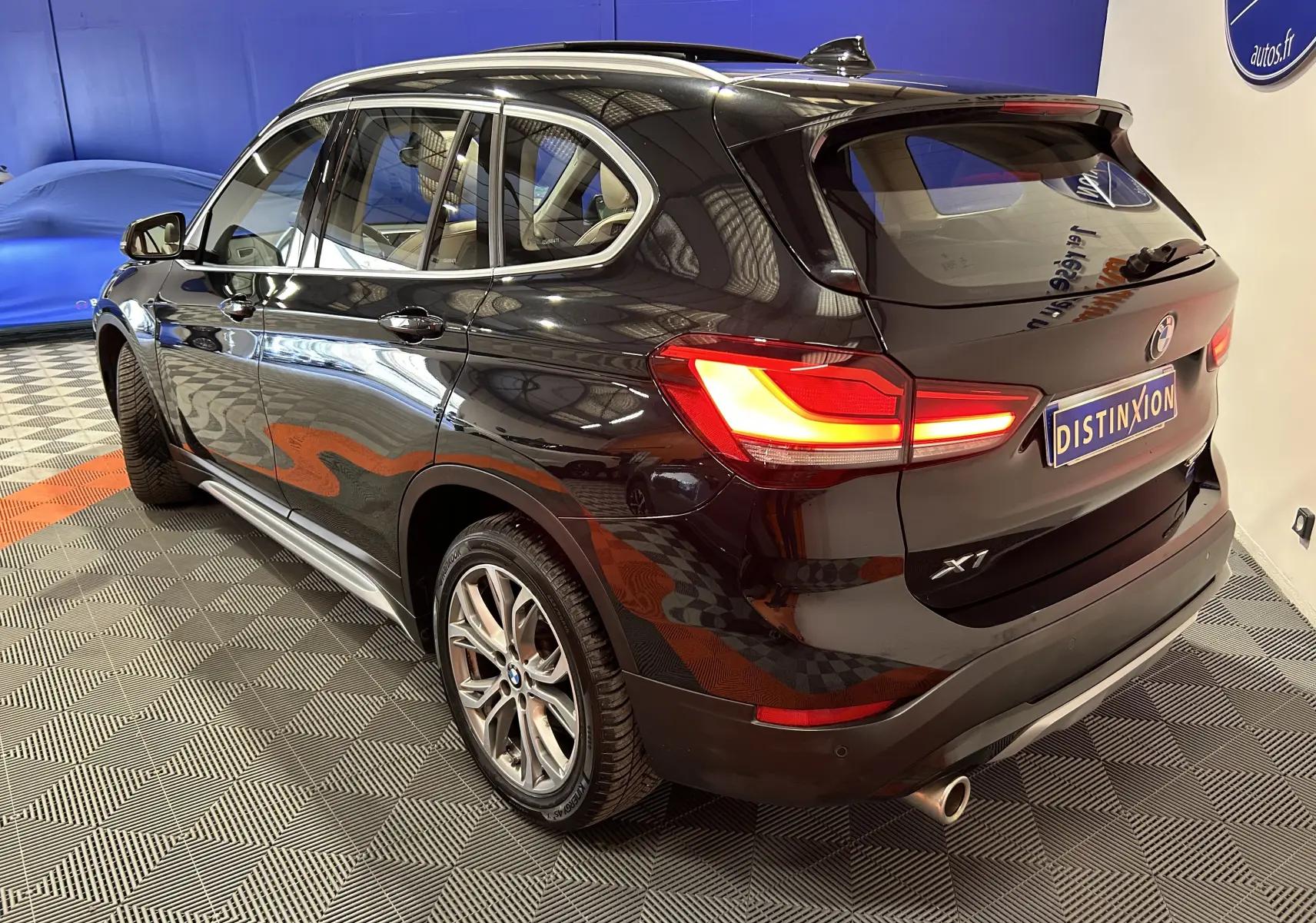 Vue 3/4 arrière droite d'un BMW X1 noir avec feux allumés et barres de toit visibles en intérieur showroom.