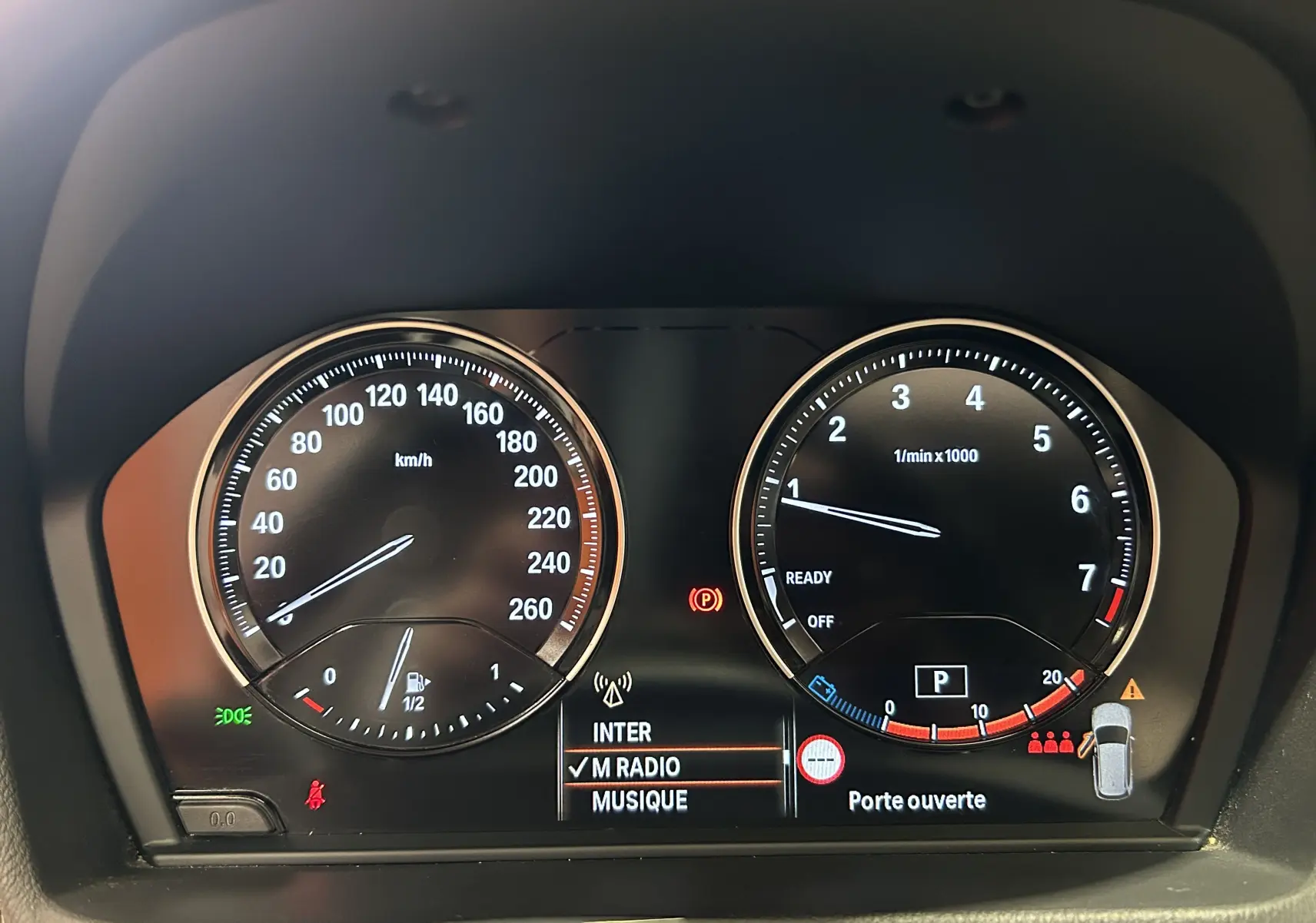 Gros plan sur le tableau de bord numérique du BMW X1 noir, affichant la vitesse, le régime moteur et un message "Porte ouverte".