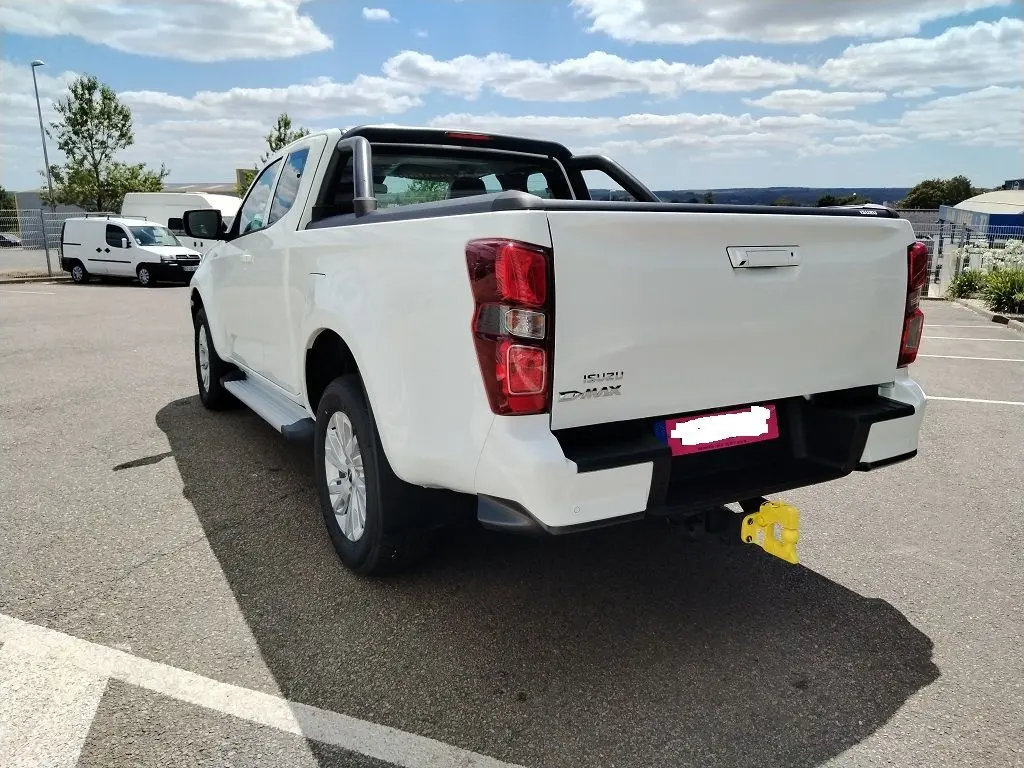 Vue 3/4 arrière droite d'un pick-up Isuzu D-MAX blanc avec arceau et volet couvre benne roulant, attelage jaune visible.