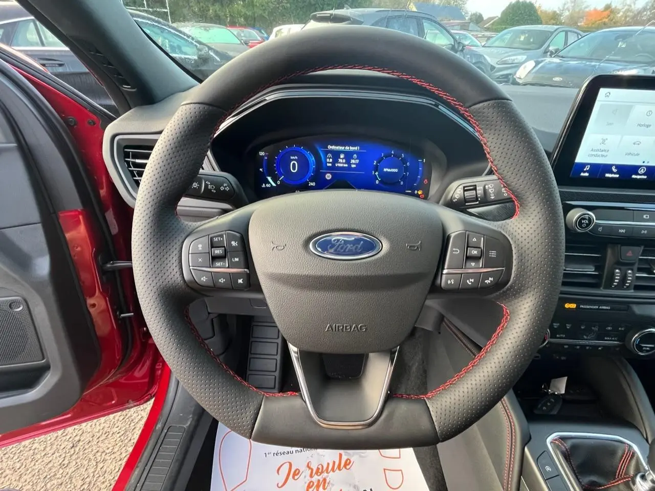 Vue centrée sur le volant perforé avec surpiqûres rouges du Ford Kuga rouge Lucifer, tableau de bord numérique et écran tactile visibles.