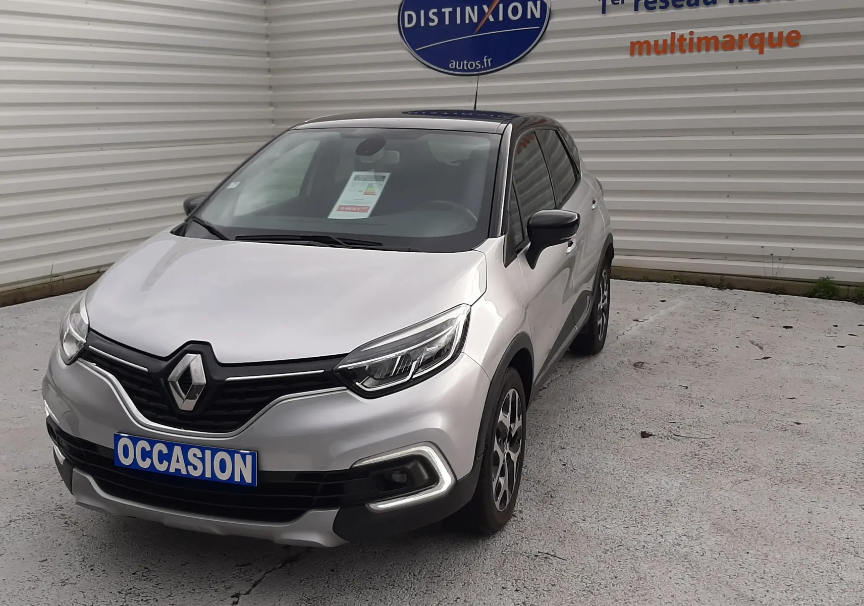 Renault Captur gris avec toit noir, vue 3/4 avant droit, stationné devant un mur blanc avec panneau Distinxion.