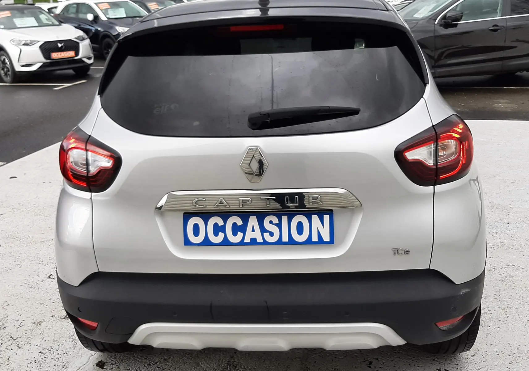 Vue arrière d'un Renault Captur gris avec toit noir, feux allumés et plaque "OCCASION" visible.