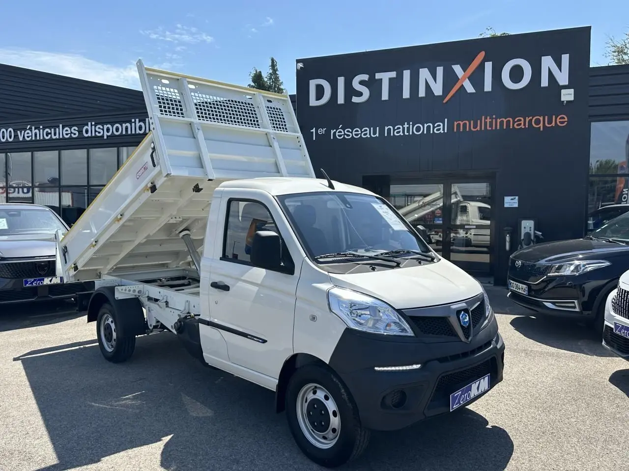 Véhicule utilitaire Piaggio Porter blanc vu en 3/4 avant droit avec benne basculante levée sur parking.