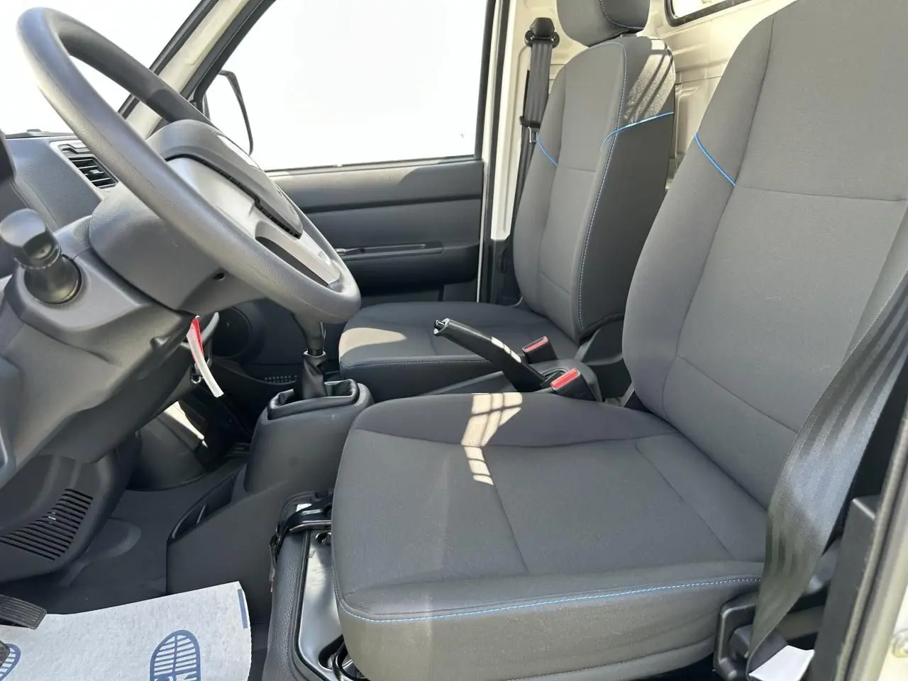 Intérieur du Piaggio Porter 2024 vu côté conducteur, sièges gris avec surpiqûres bleues et volant noir.