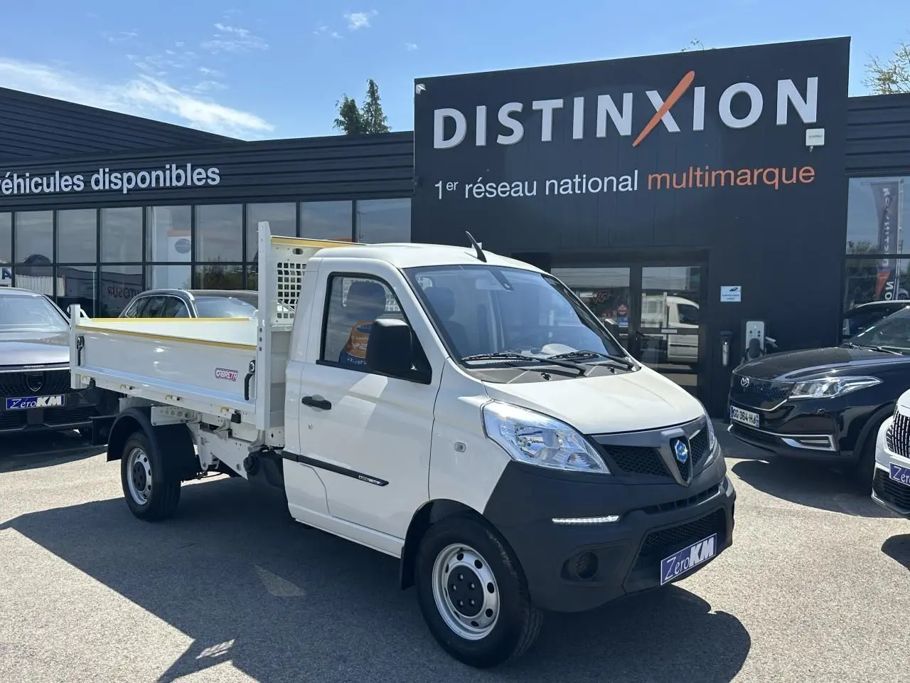 Vue 3/4 avant droite d'un Piaggio Porter blanc avec benne longue, stationné devant un concessionnaire multimarque.