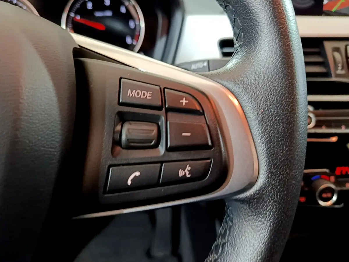 Gros plan sur les commandes gauche du volant cuir noir du BMW X1 2021, avec tableau de bord en arrière-plan.