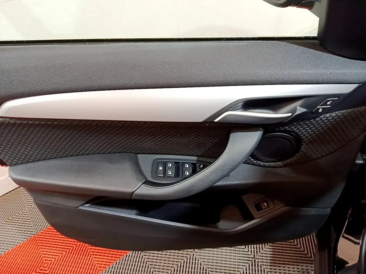 Vue intérieure côté gauche de la porte avant d'une BMW X1 noire, avec garniture carbone et commandes de vitres.