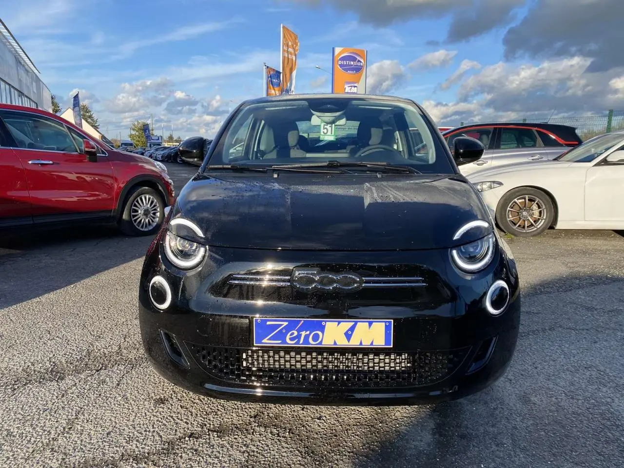 Vue avant d'une FIAT 500 électrique noire avec phares ronds LED et plaque Zéro KM sur un parking.