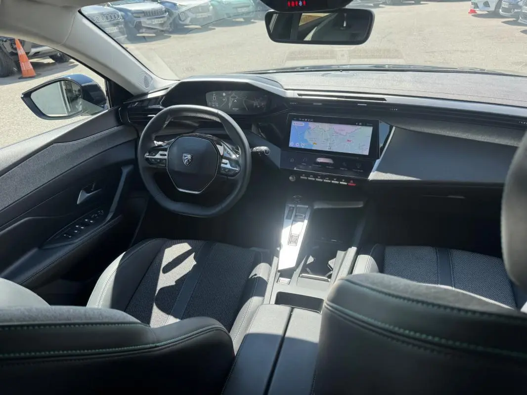 Vue intérieure avant de la Peugeot 308 Hybrid noire, montrant le volant, le cockpit numérique et l'écran central tactile avec navigation.