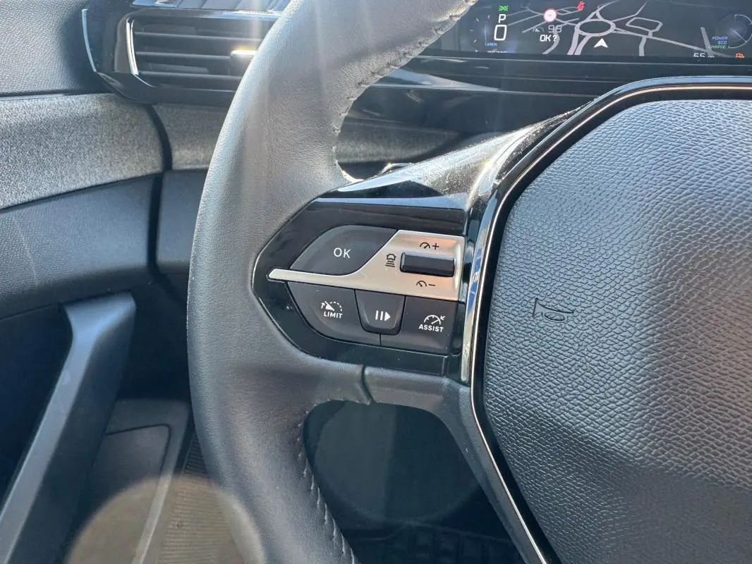 Gros plan sur le volant noir de la Peugeot 308 Hybrid 2024, avec commandes intégrées et cockpit numérique en arrière-plan.