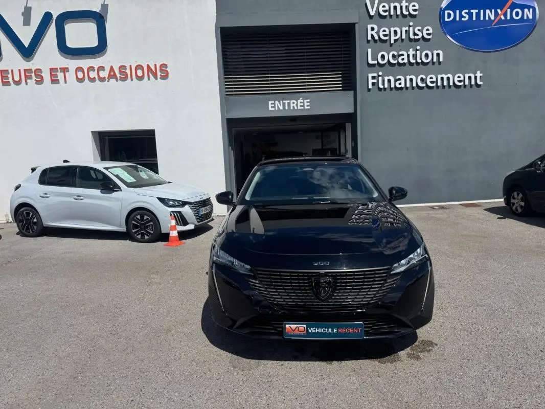 Vue frontale d'une Peugeot 308 Hybrid 136 e-DCS6 Allure noire, garée devant une concession avec reflet du ciel sur la carrosserie.