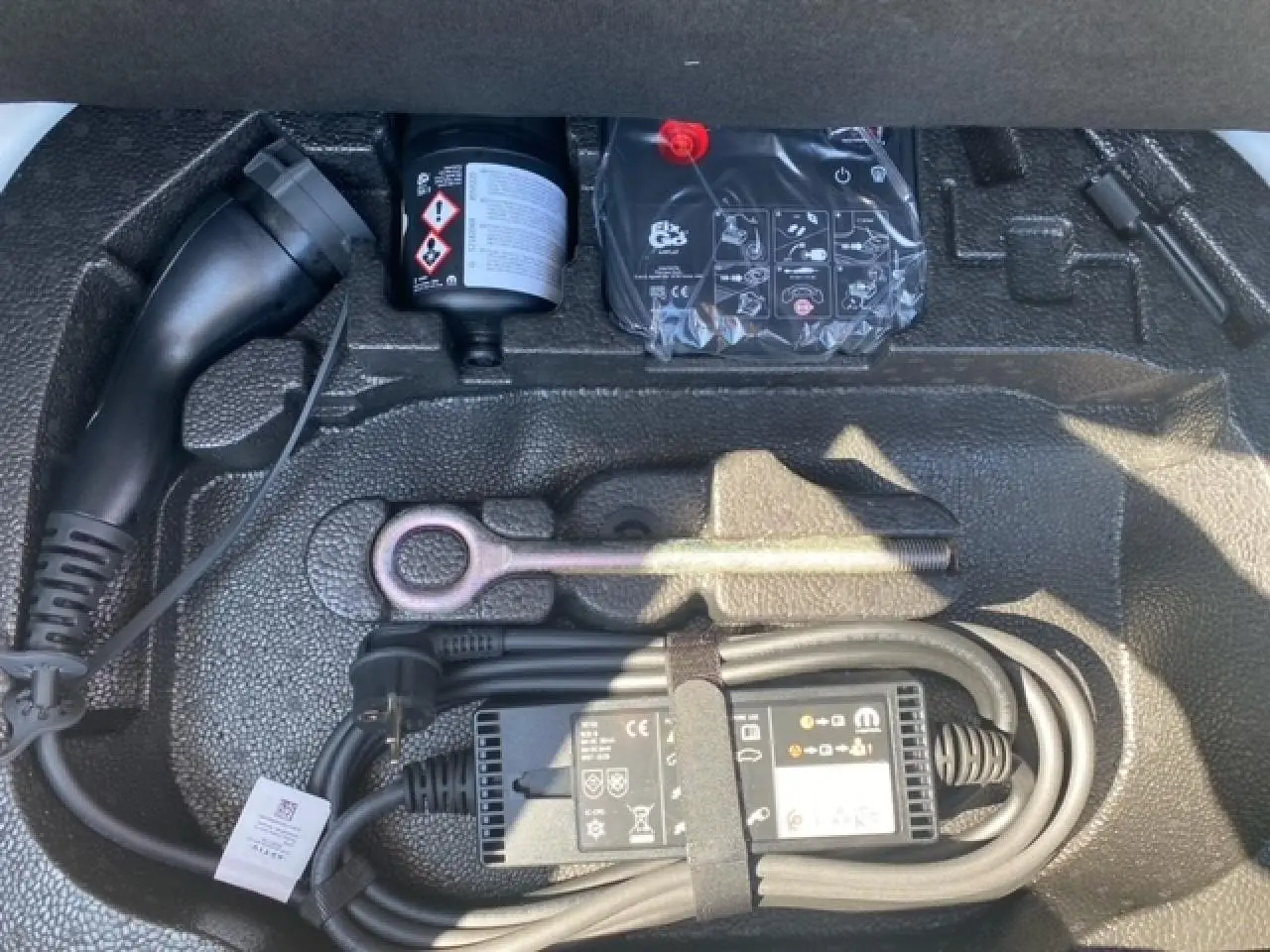 Gros plan sur le compartiment de rangement sous coffre d'une FIAT 500 électrique blanche avec câble de recharge et outils.