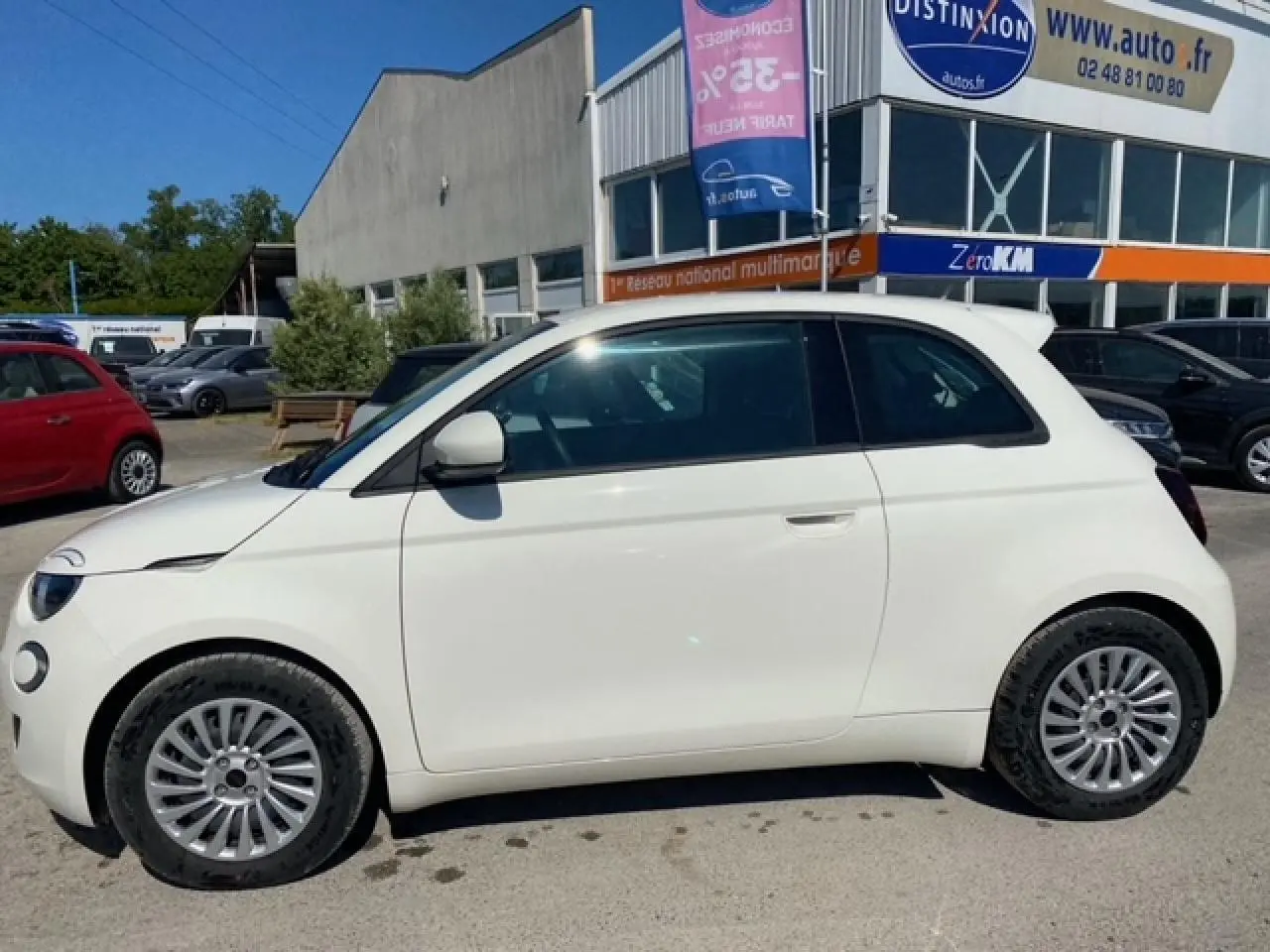 Profil côté gauche d'une FIAT 500 électrique blanche Ice White, avec jantes alliage et toit noir en arrière-plan d'un parking.