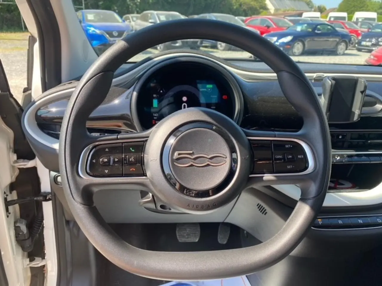 Vue intérieure centrée sur le volant noir de la Fiat 500 électrique blanche avec tableau de bord numérique visible.