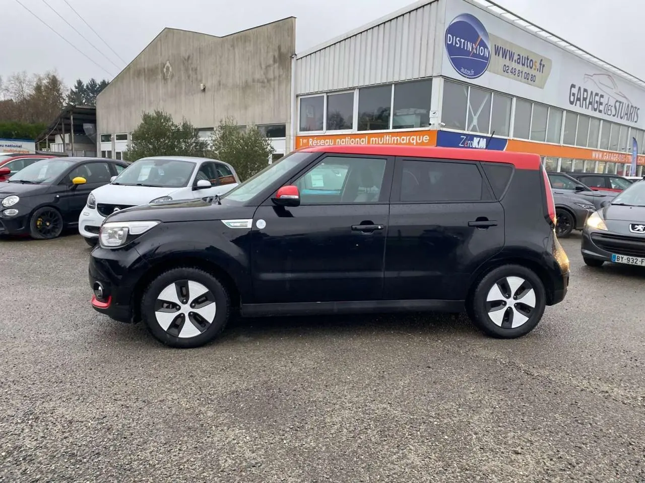 KIA SOUL électrique noir avec toit rouge vu de profil côté gauche sur un parking devant un garage multimarque.