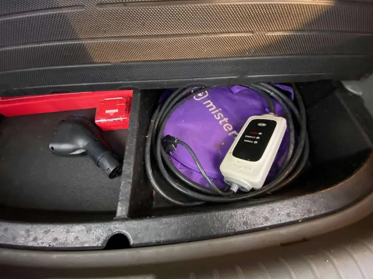 Gros plan sur le compartiment de rangement avant d'une KIA Soul électrique 2016, montrant un câble de recharge et une trousse violette.