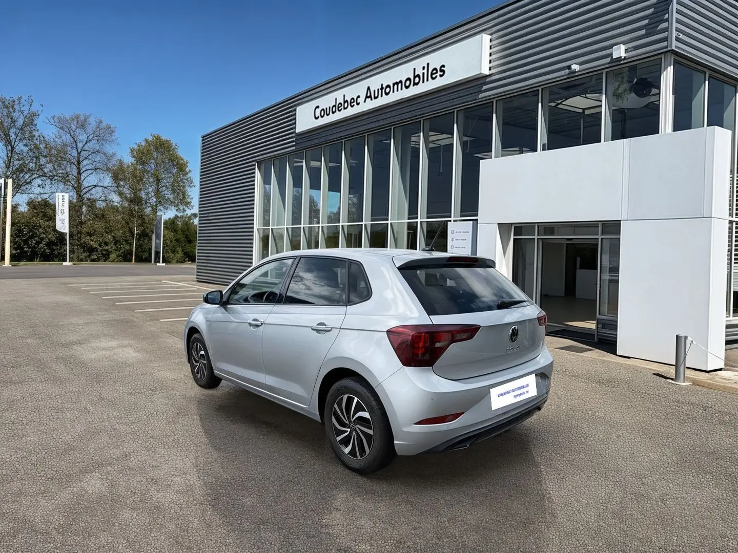 Vue 3/4 arrière droite d'une Volkswagen Polo grise 2025 stationnée devant un bâtiment de concession.
