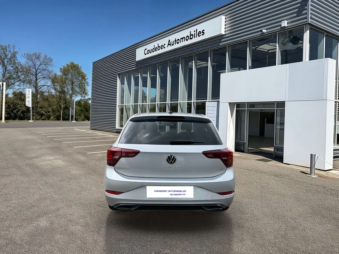 Vue arrière d'une Volkswagen Polo grise 2025 garée devant un bâtiment moderne de concession automobile.