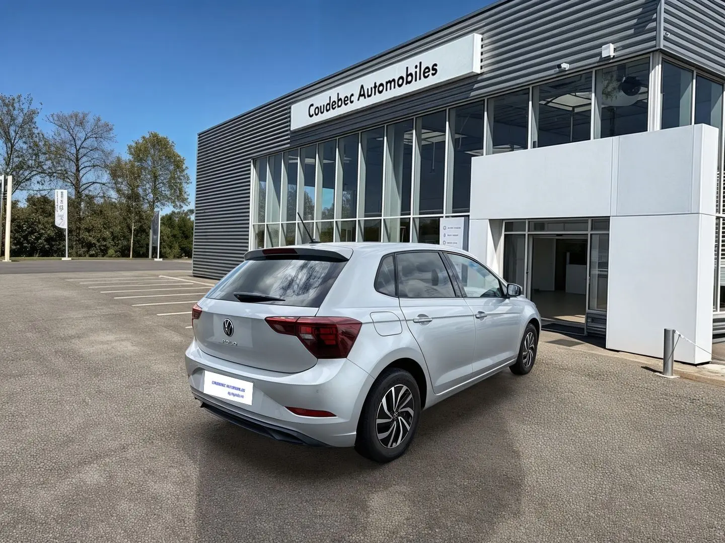 Vue 3/4 arrière droite d'une Volkswagen Polo grise stationnée devant un bâtiment moderne de concession automobile.