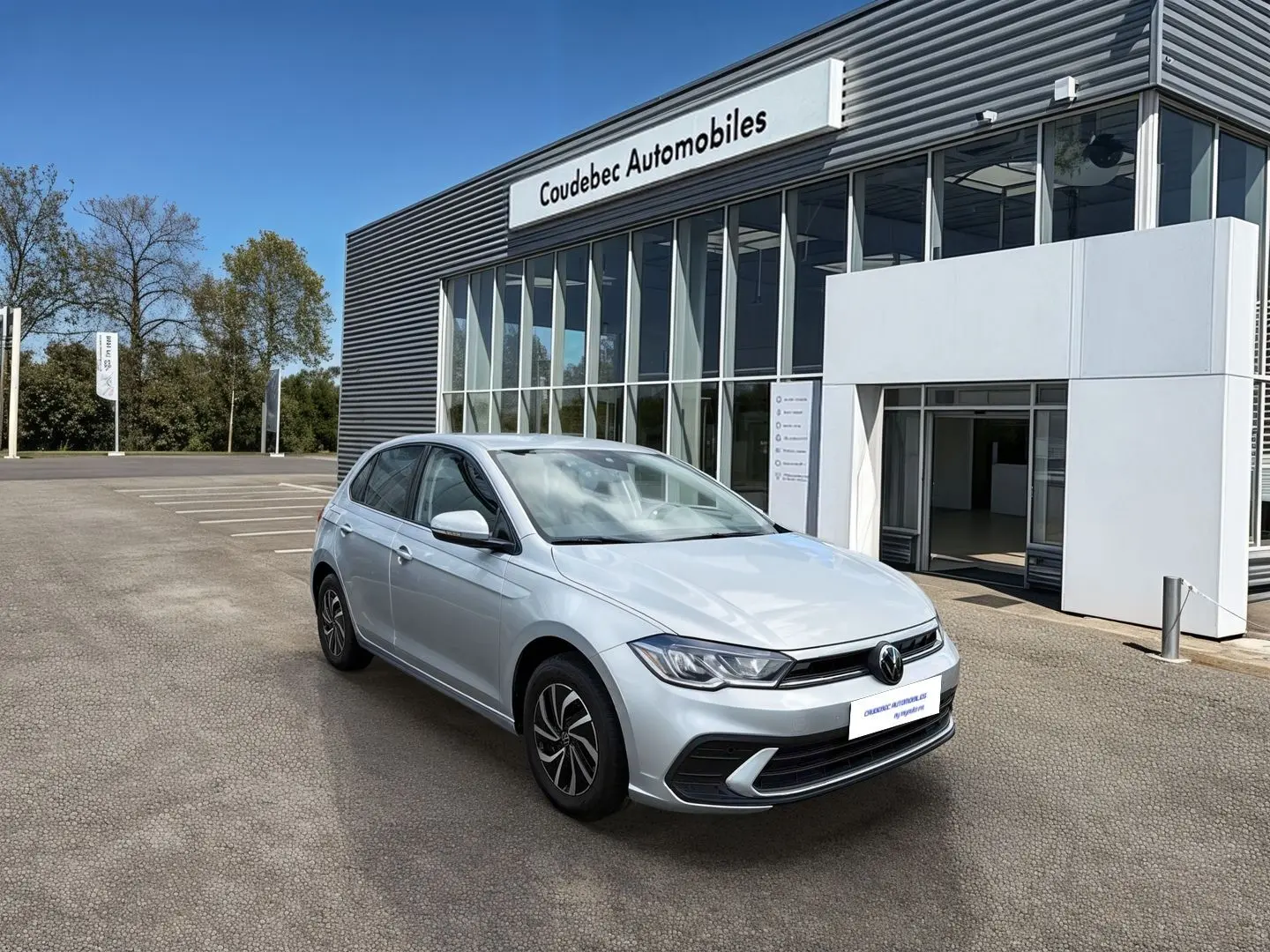 Volkswagen Polo grise vue 3/4 avant droit devant un showroom Coudebec Automobiles, feux LED allumés.