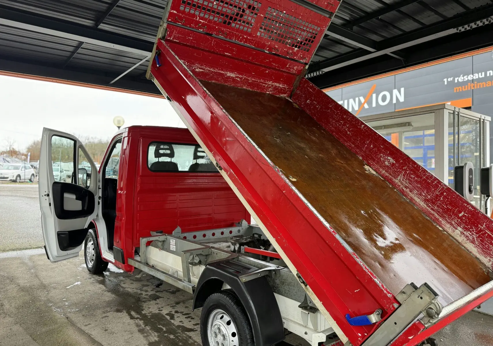 Vue 3/4 arrière gauche d'un Citroën Jumper benne rouge avec benne relevée et porte conducteur ouverte.