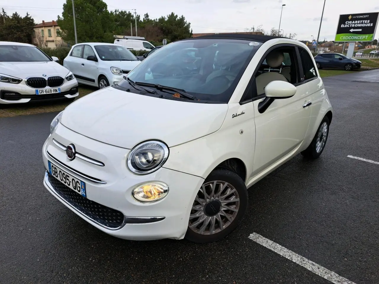 Fiat 500 Cabriolet blanche vue en 3/4 avant droit, toit ouvert partiellement, phare avant droit allumé sur parking.