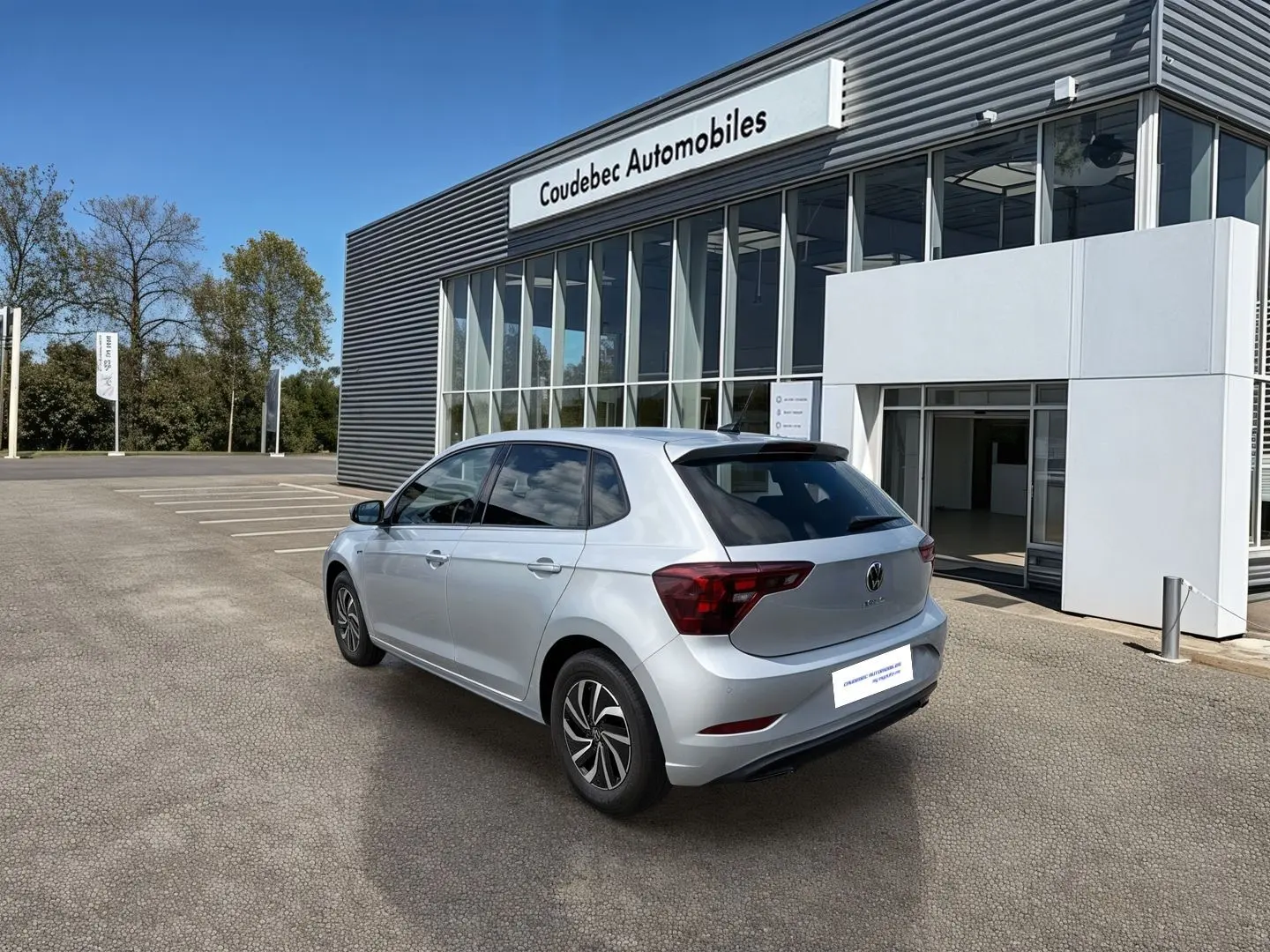 Vue 3/4 arrière droite d'une Volkswagen Polo grise stationnée devant un bâtiment moderne de concession automobile.