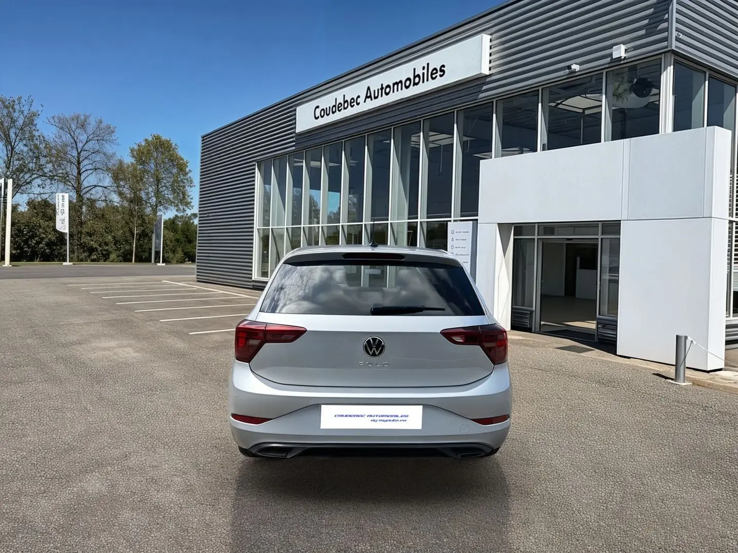 Vue arrière d'une Volkswagen Polo grise 2025 garée devant un bâtiment moderne de concession automobile.