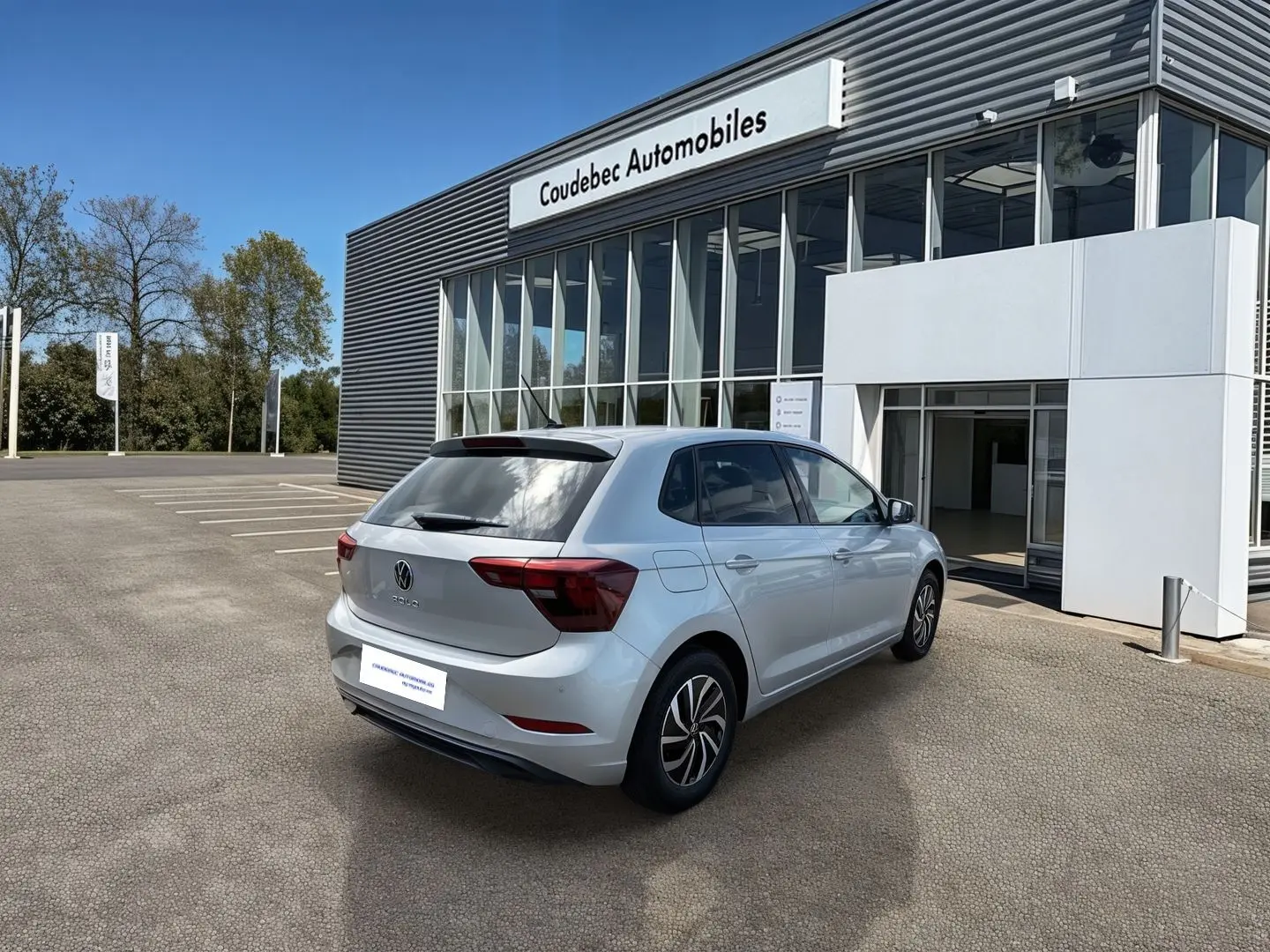 Volkswagen Polo grise vue 3/4 arrière droit, stationnée devant un bâtiment de concession Coudebec Automobiles.