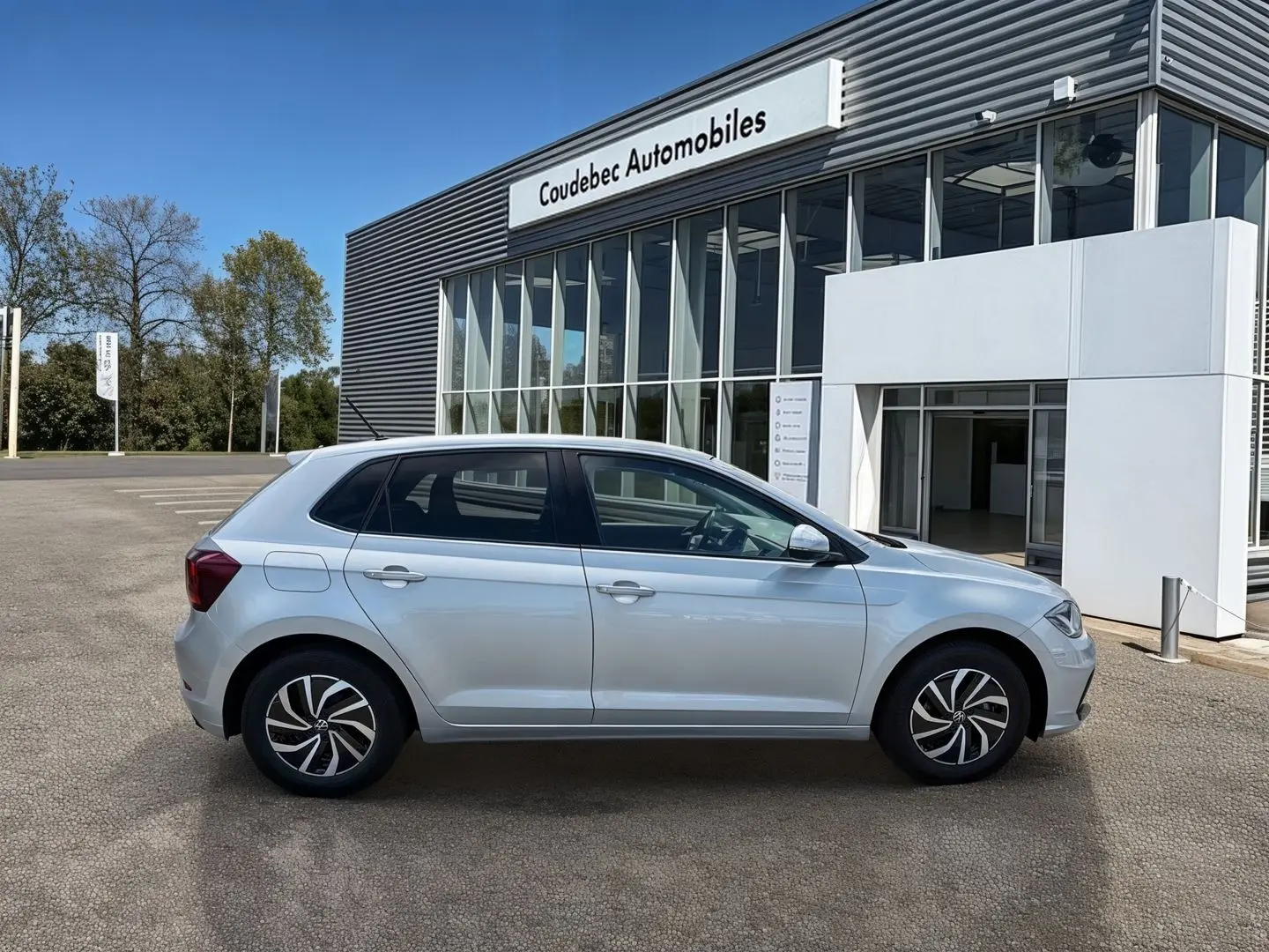 Profil côté gauche d'une Volkswagen Polo grise 2025 garée devant un bâtiment moderne de concession automobile.