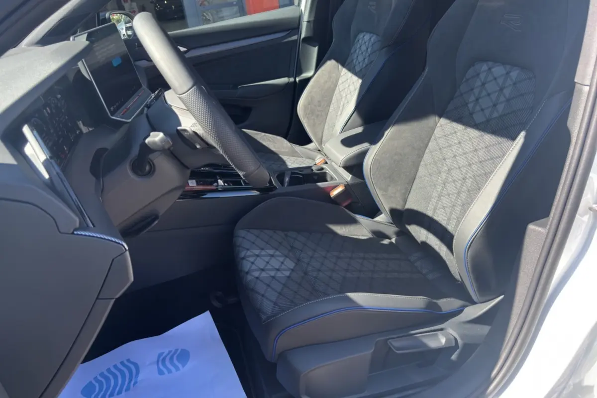 Vue intérieure côté conducteur de la Volkswagen Golf TDI 150 R-Line 2026, sièges noirs avec surpiqûres bleues et volant cuir multifonctions.