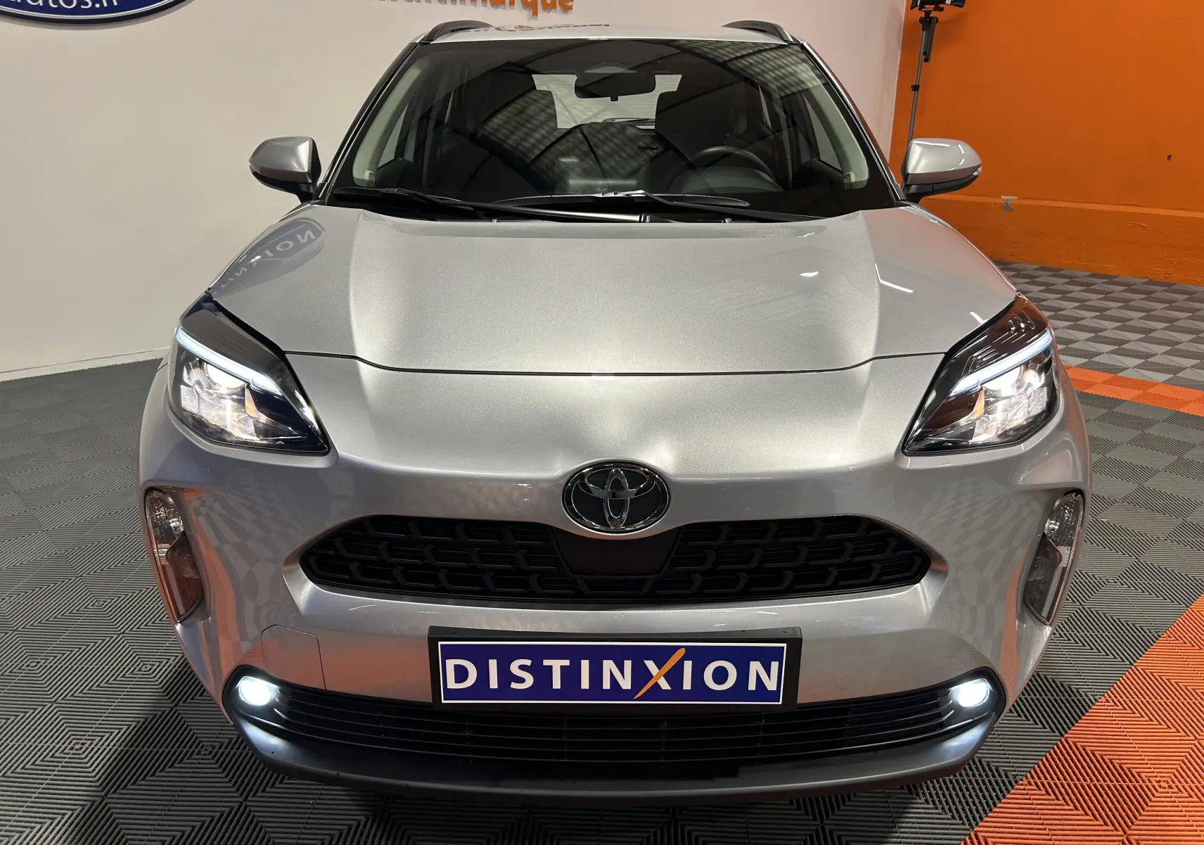 Vue avant d'une Toyota Yaris Cross 116h Active gris minéral avec phares allumés et plaque Distinxion en intérieur showroom.