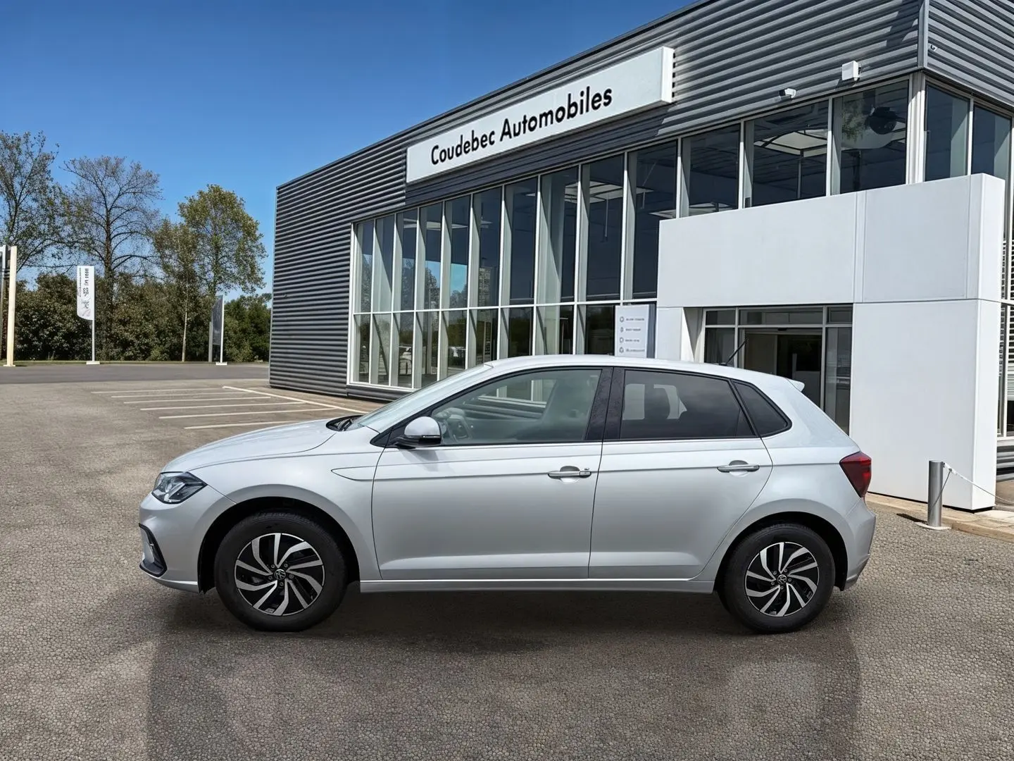 Vue de profil côté gauche d'une Volkswagen Polo grise stationnée devant un bâtiment commercial moderne.
