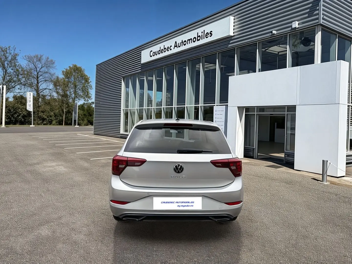 Vue arrière d'une Volkswagen Polo grise 2025 stationnée devant un bâtiment moderne de concession automobile.