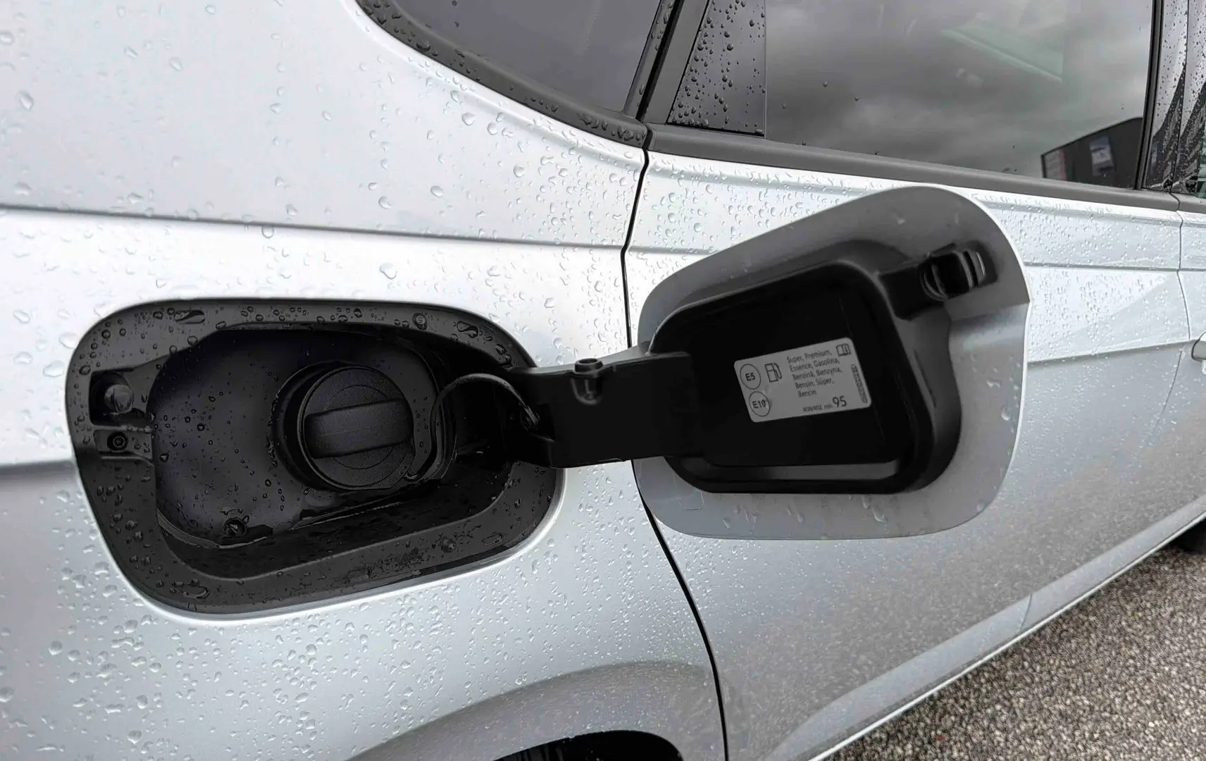 Gros plan sur la trappe à carburant ouverte côté gauche d'une Volkswagen Polo grise sous la pluie.