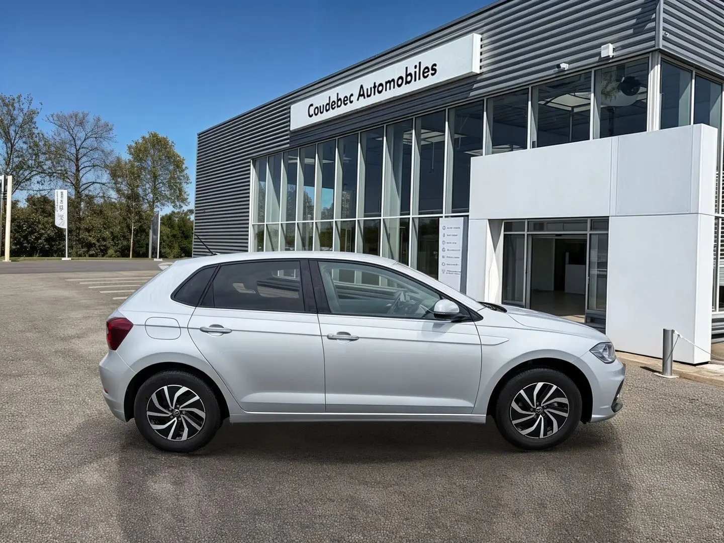 Vue de profil côté gauche d'une Volkswagen Polo grise 2025 stationnée devant un bâtiment commercial.