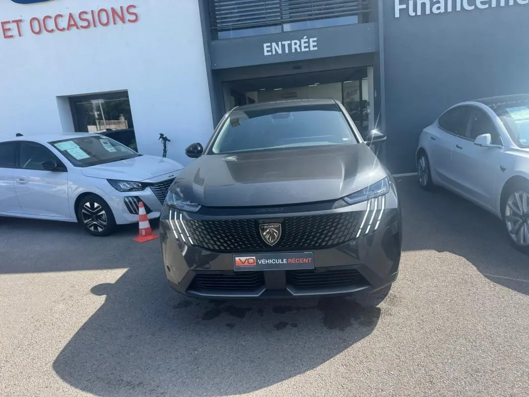 Vue avant d’un Peugeot 3008 Hybrid gris foncé avec calandre noire et feux LED en forme de griffes, stationné devant un bâtiment.
