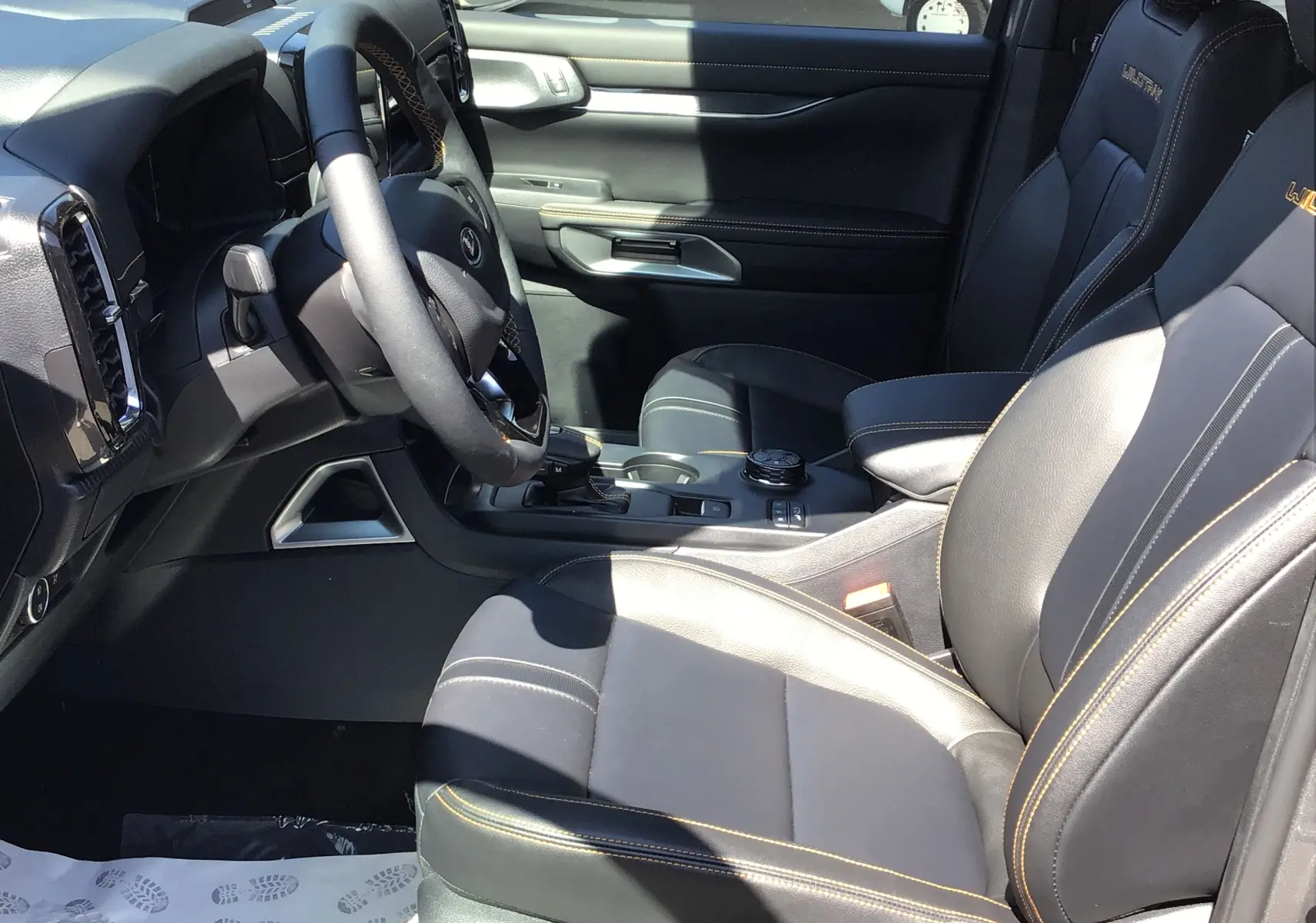 Vue intérieure côté conducteur du Ford Ranger Wildtrak gris carbone, sièges cuir noirs avec surpiqûres orange et volant multifonction.