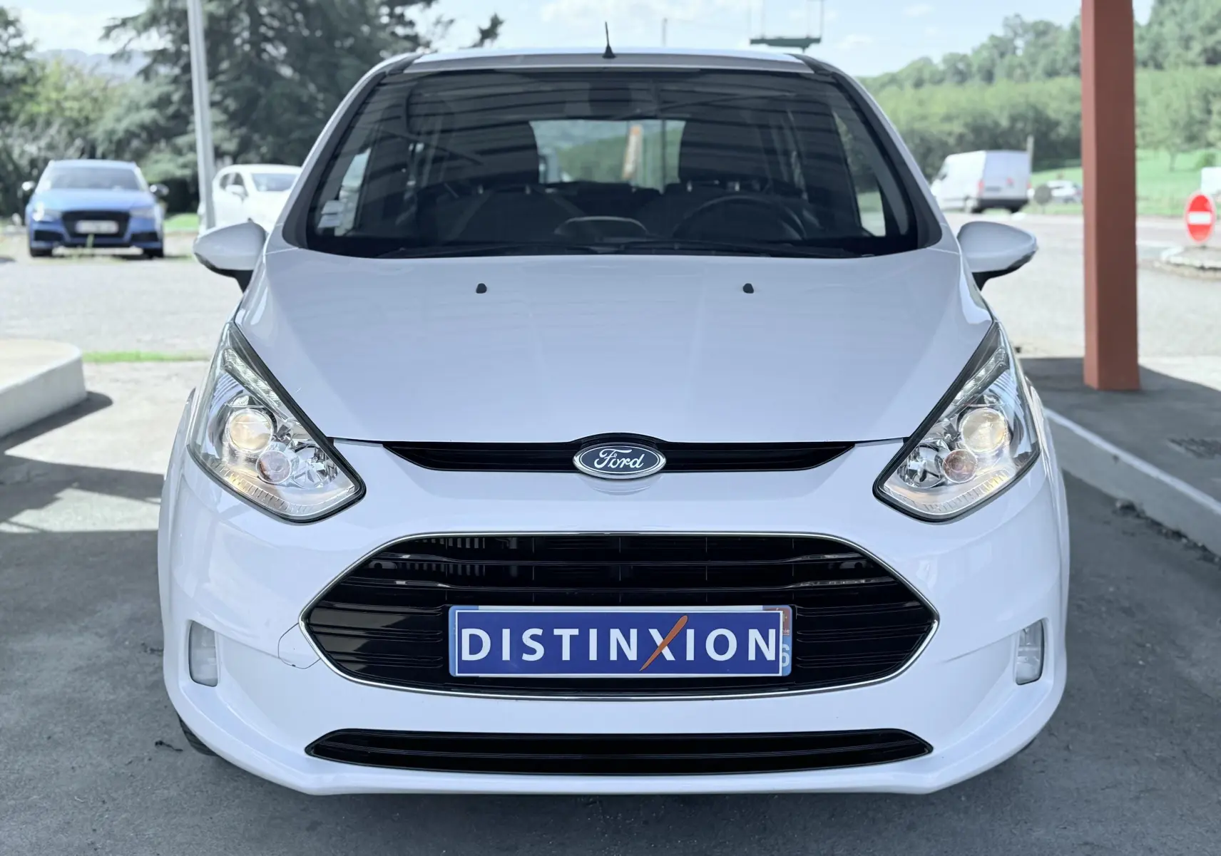 Vue frontale d'une Ford B-MAX blanche 2017 avec calandre noire et phares allumés, plaque Distinxion visible.