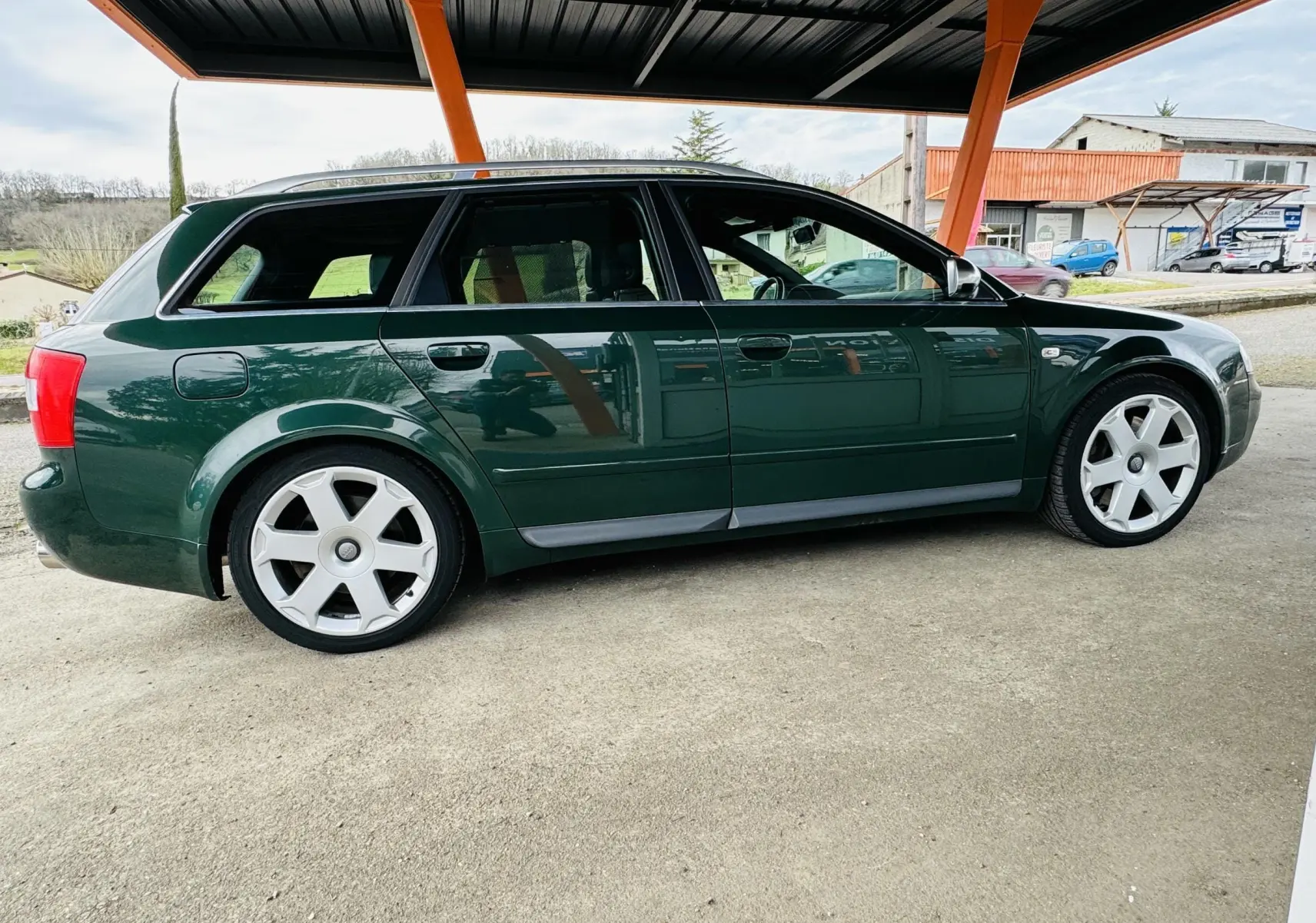 Profil latéral droit de l'Audi S4 Avant 2004 vert foncé avec jantes alliage distinctives sous un abri métallique orange.